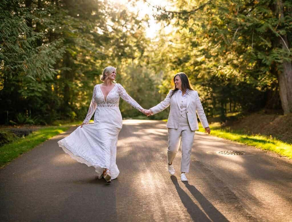 Two brides walk down the treelined street hand in hand on their wedding day